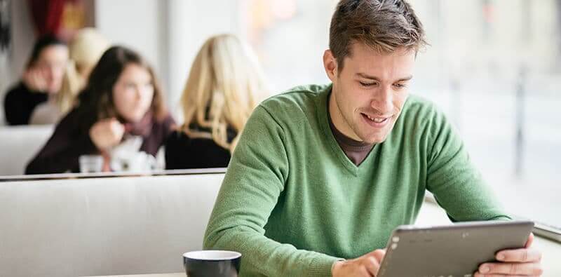Mand i grøn sweater sidder og kigger på sin tablet med en kop kaffe og smiler
